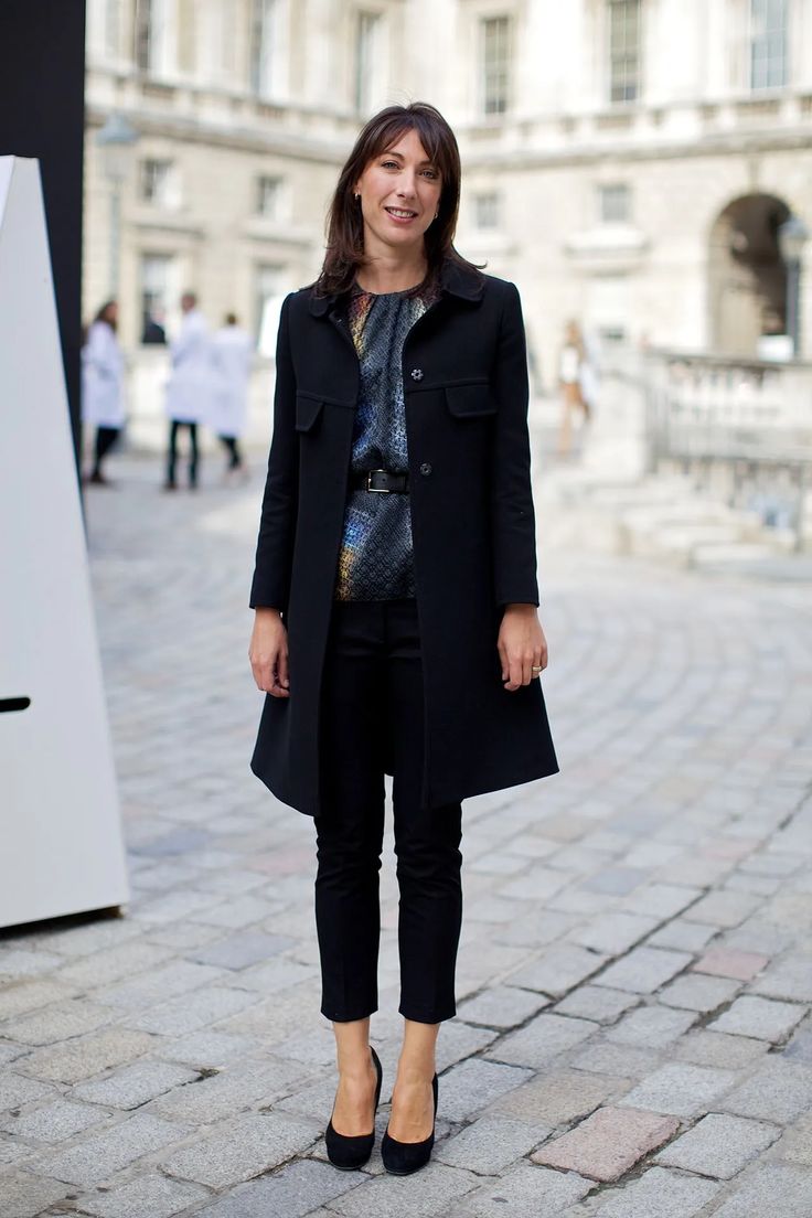 a woman standing in front of a building wearing black pants and a coat with buttons on it