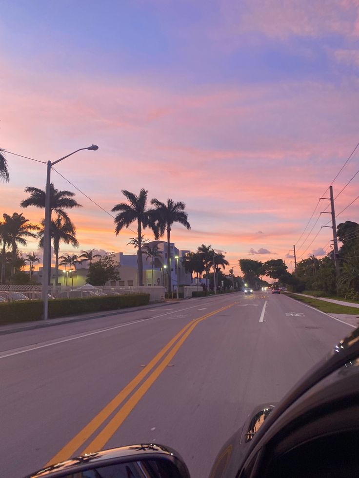 the sun is setting behind palm trees on the side of the road as cars drive down the street