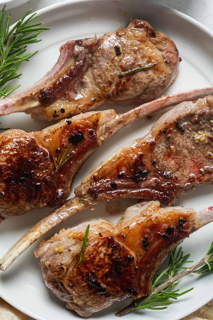 three pieces of meat on a white plate with rosemary sprigs and seasoning