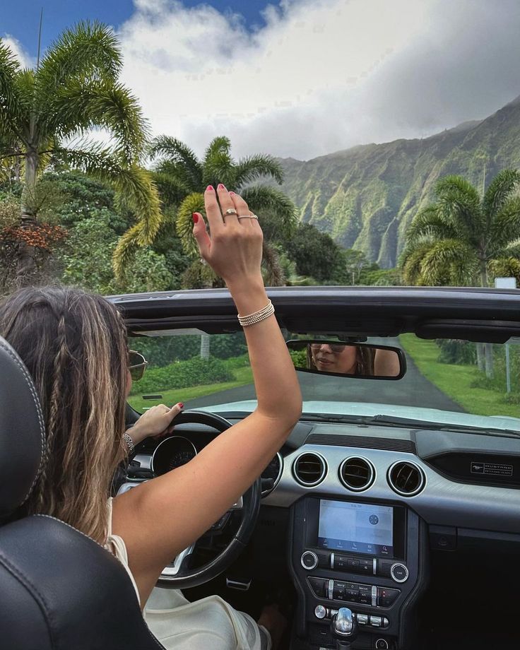 a woman driving a car with her hand up in the air and palm trees behind her