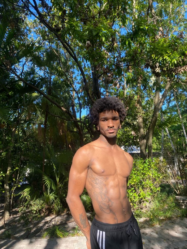 a shirtless young man standing in front of some trees and bushes with his hands on his hips