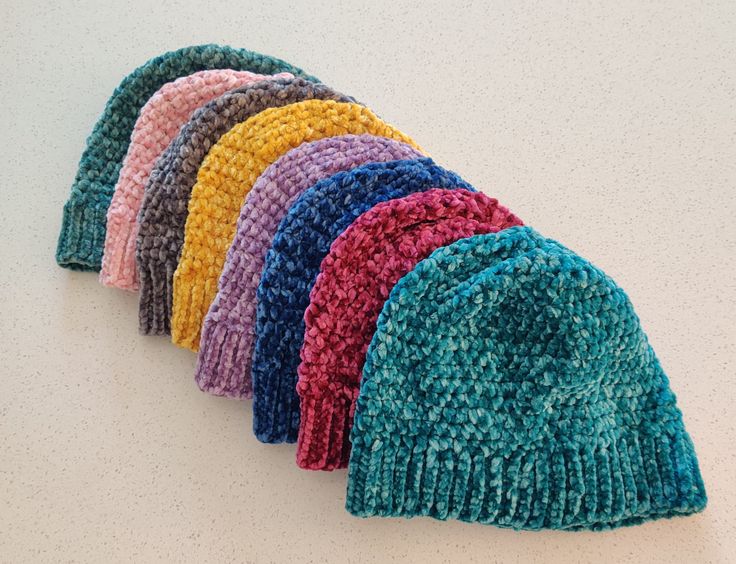 six knitted hats lined up on a white counter top, all in different colors