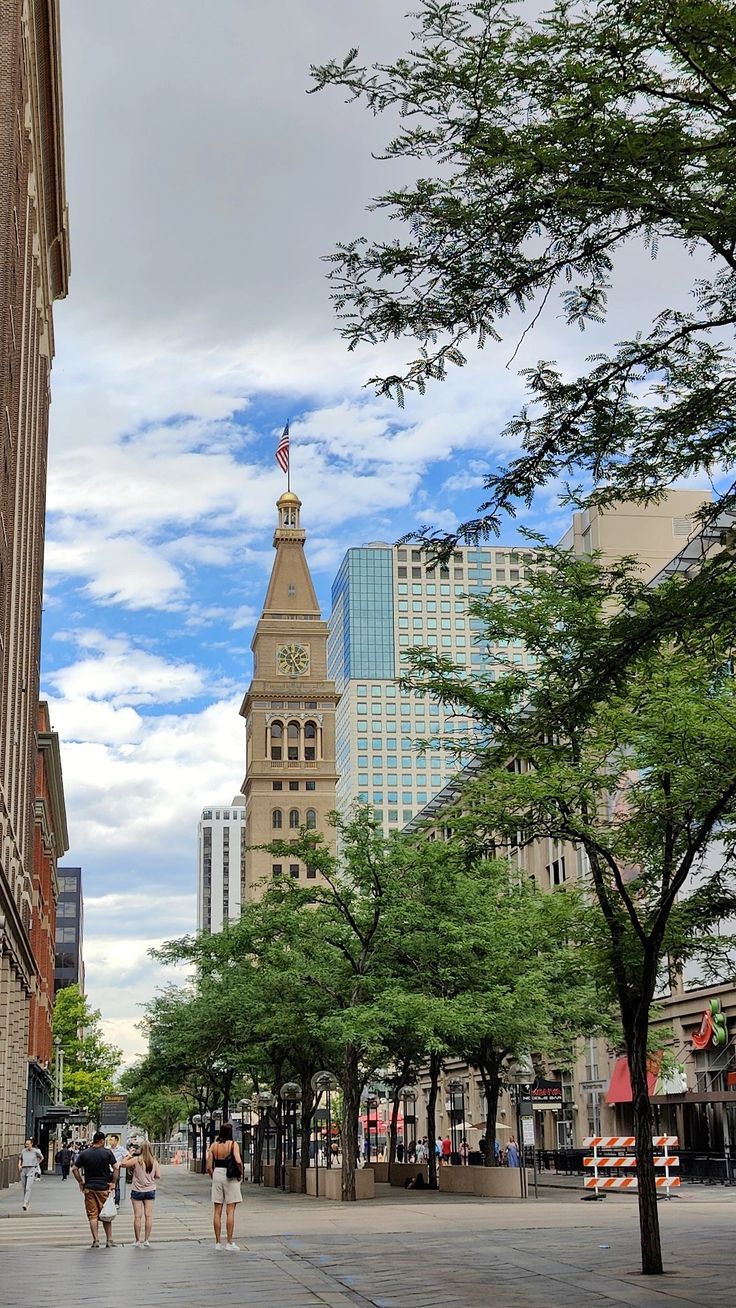 two people walking down the street in front of tall buildings with trees on both sides