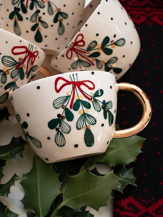 white cups with red bows and holly on them are sitting next to some green leaves