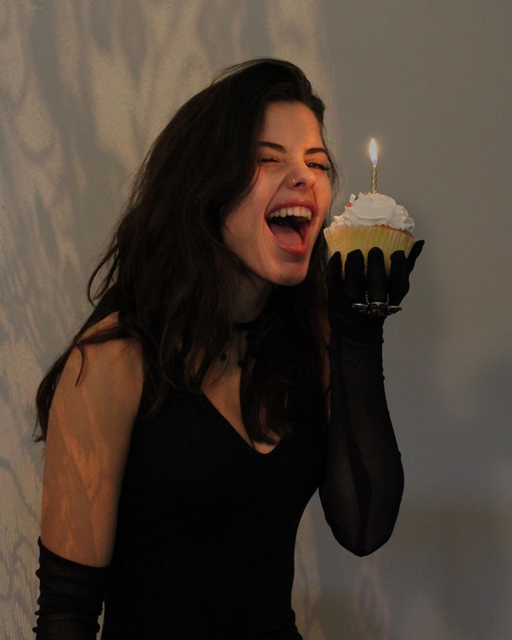 a woman holding up a cupcake with a candle in it's mouth and wearing black gloves