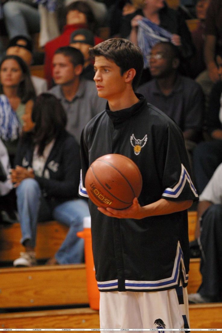 a young man holding a basketball while standing on a court with people watching from the bleachers