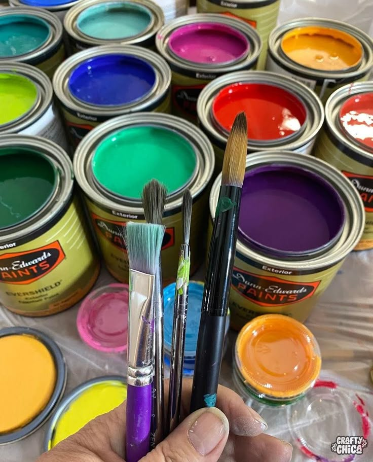 a person holding some paint and brushes in their hand next to many different color paints