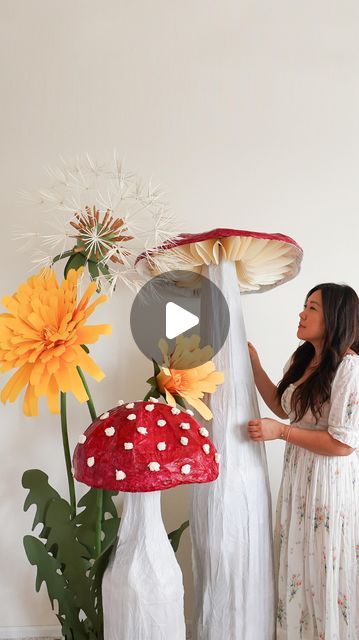 a woman standing in front of a mushroom sculpture with flowers behind her and an orange flower next to it