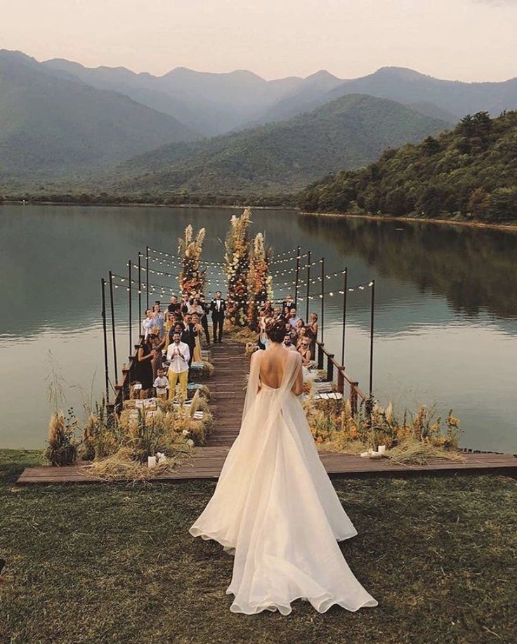 a woman in a wedding dress is standing on the edge of a dock with mountains in the background