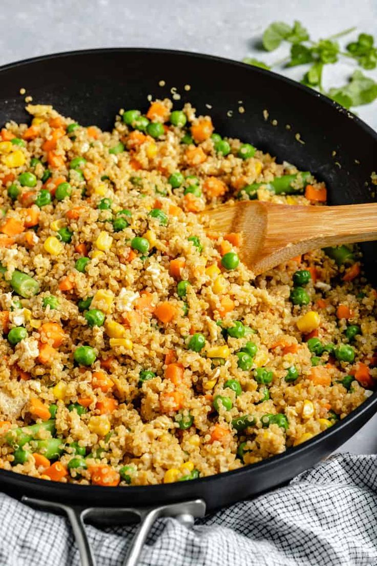 a skillet filled with rice and vegetables on top of a cloth next to a wooden spoon