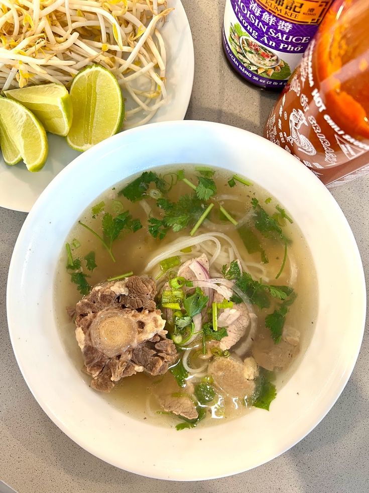 a bowl of pho noodle soup with limes on the side and a bottle of beer next to it