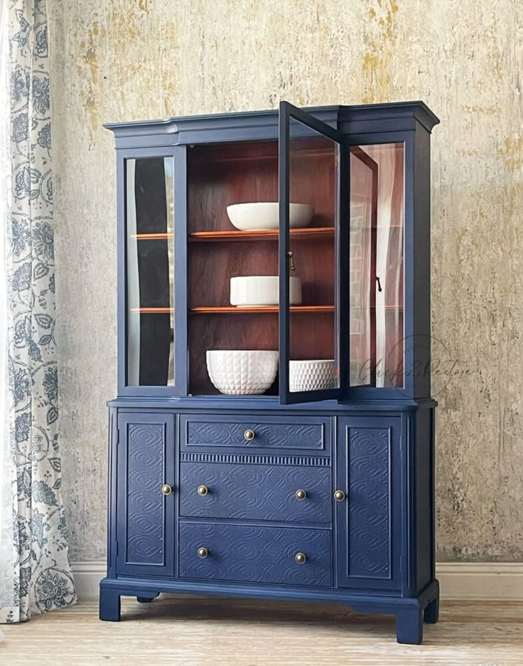 a blue china cabinet with glass doors and drawers on the bottom, in front of a beige wall