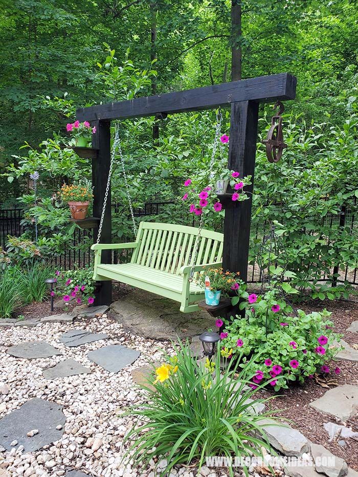 a green bench sitting in the middle of a garden next to flowers and trees with hanging planters