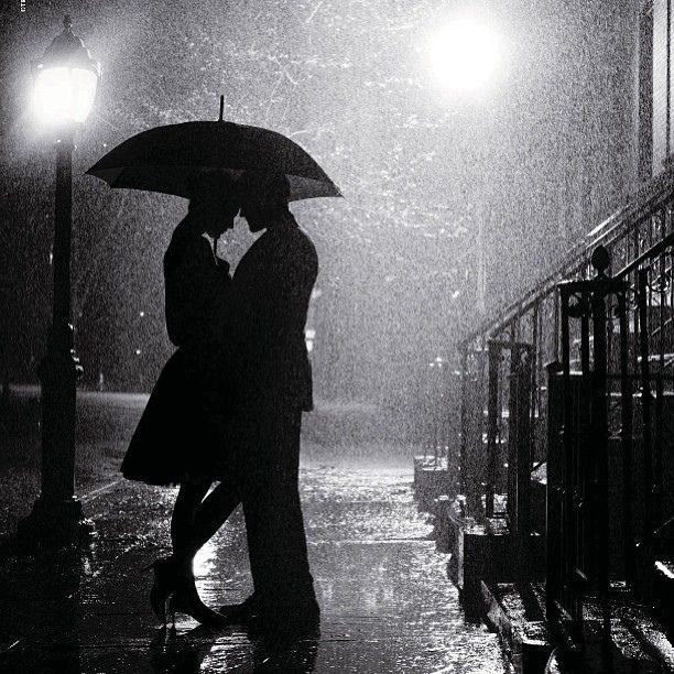 a couple kissing under an umbrella on a rainy night in the rain, with street lights lit up behind them