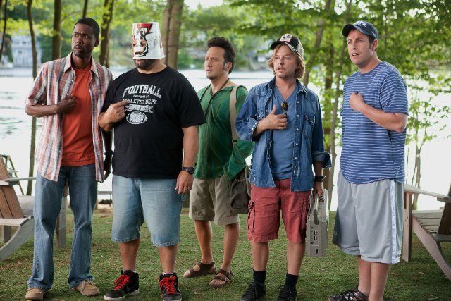 four men standing in front of a lake with trees and grass on the ground, one wearing a paper hat