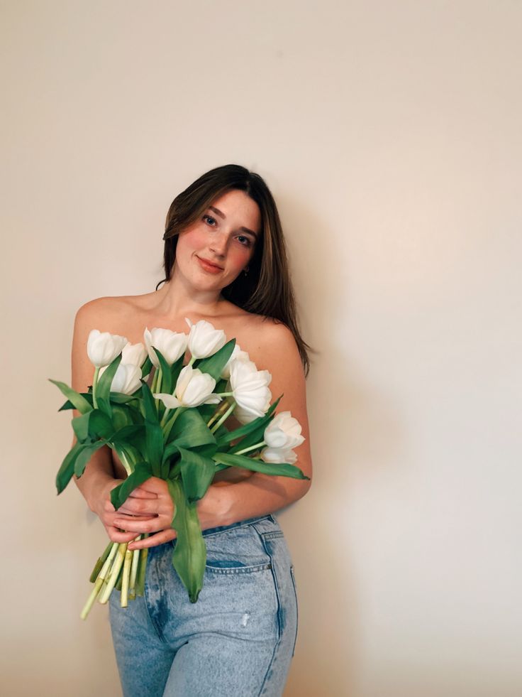 a woman holding flowers in her hands and looking at the camera while standing against a wall