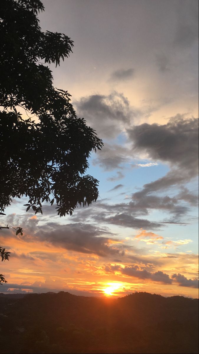 the sun is setting behind some trees and hills in the distance, with clouds above it