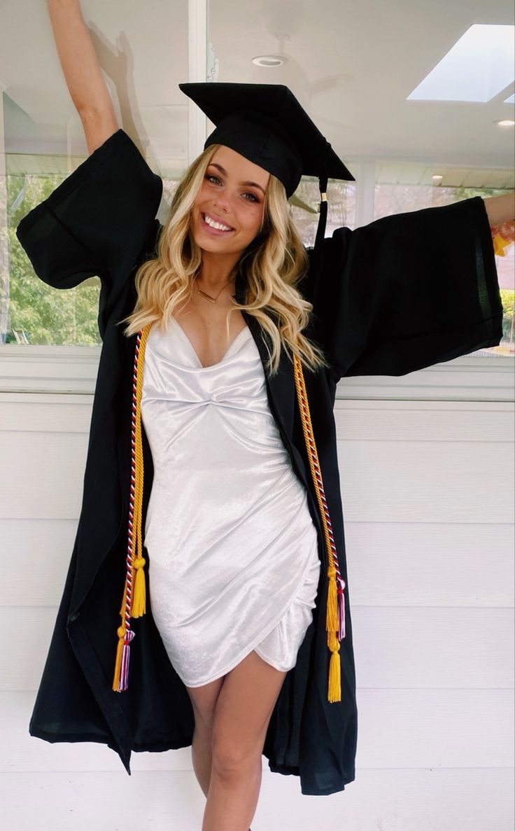 a woman wearing a graduation gown and holding her arms in the air while standing next to a window