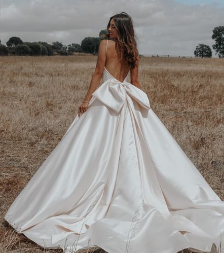 a woman standing in a field wearing a wedding dress with a bow on the back