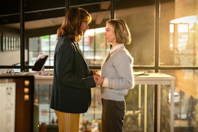 two women standing in front of a counter talking to each other with sunlight streaming through the window behind them