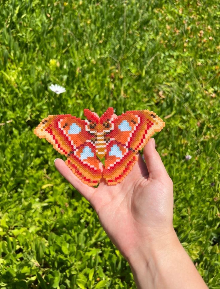 a hand holding an orange beaded butterfly in front of some green grass and bushes