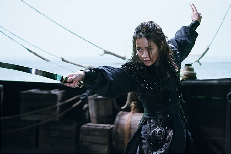 a woman dressed in black is holding her arms out while standing on the deck of a ship