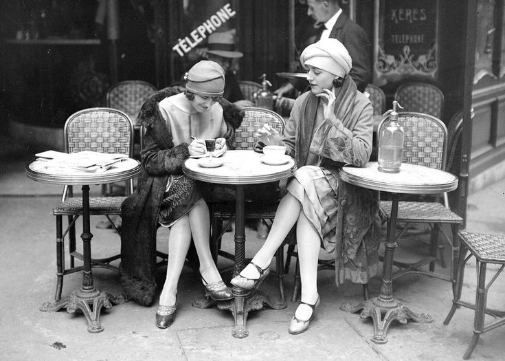 two women sitting at an outdoor table talking on their cell phones in front of a store