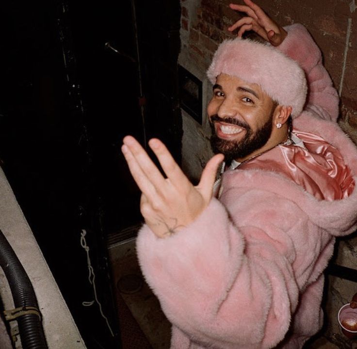 a man wearing a pink fur coat and holding his hand up in the air while standing next to a brick wall