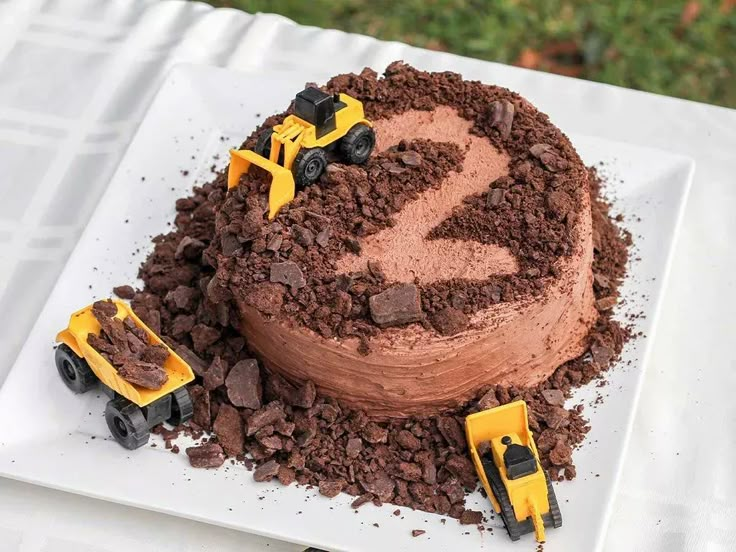 a chocolate cake with construction trucks on top is sitting on a white platter and has dirt around it