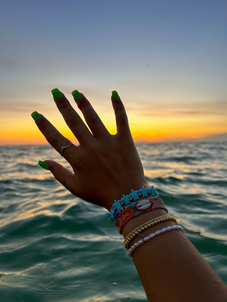 a woman's hand with green nails and bracelets on the water at sunset