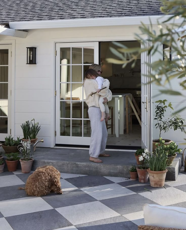 a woman holding a baby standing in front of a house with potted plants on the porch