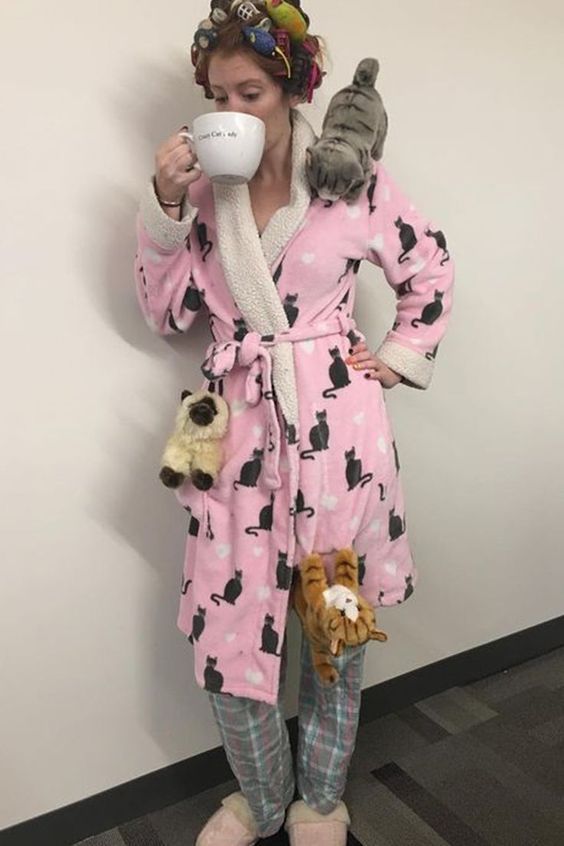 a woman in pink robe holding a coffee cup and two cats on her lap while standing next to a wall