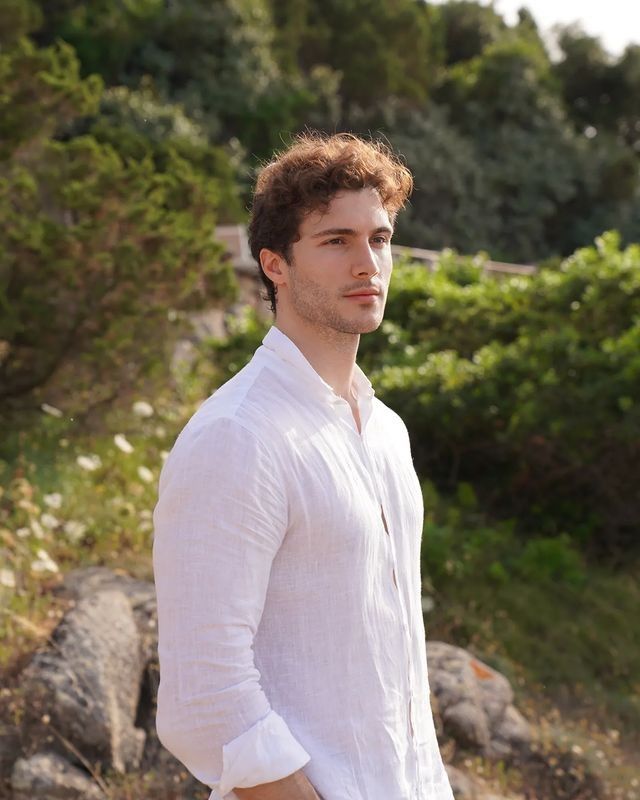 a man in white shirt and jeans standing by some rocks with his hands in his pockets