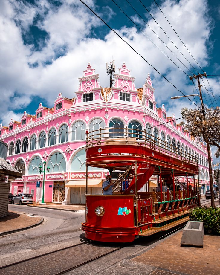 a red double decker bus driving past a pink and white building with lots of windows