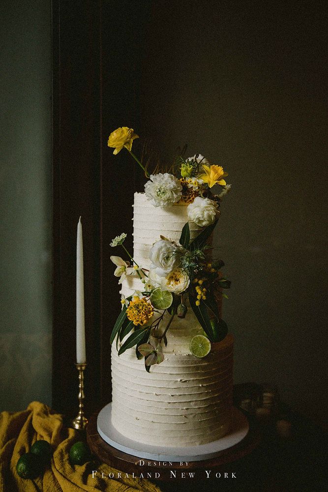 a white wedding cake with yellow flowers and greenery next to a candle on a table