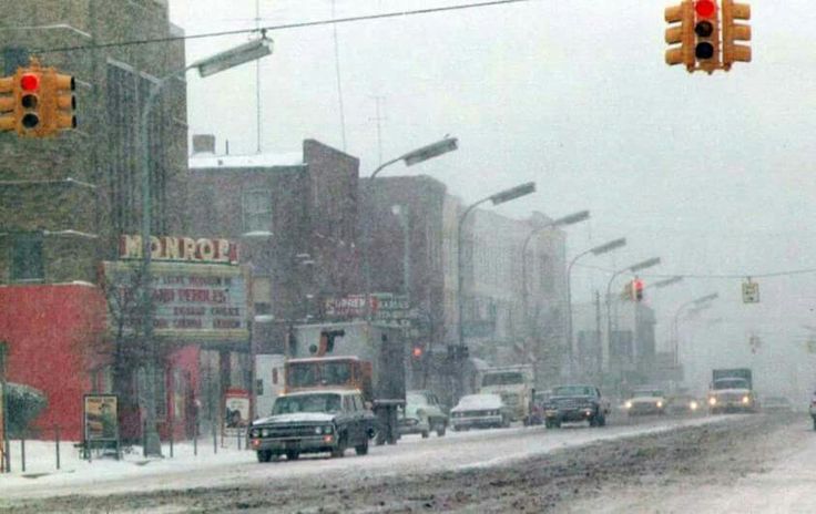 cars driving down a snow covered street with traffic lights and buildings in the background on a snowy day