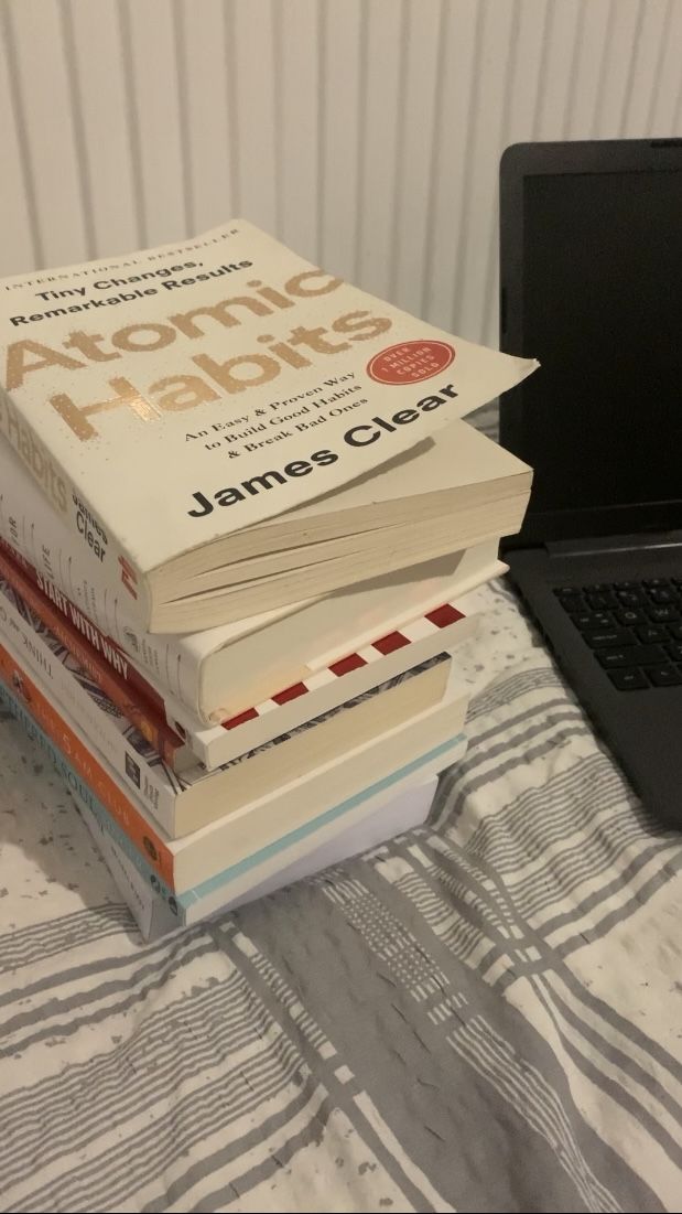 a stack of books sitting next to a laptop computer on top of a bed in front of a wall