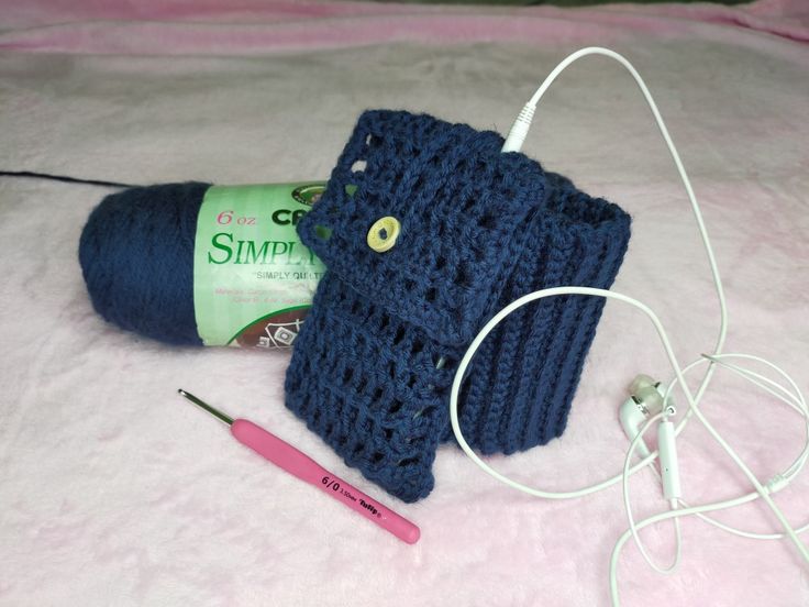 a blue crocheted cell phone case sitting on top of a bed next to a pair of ear buds