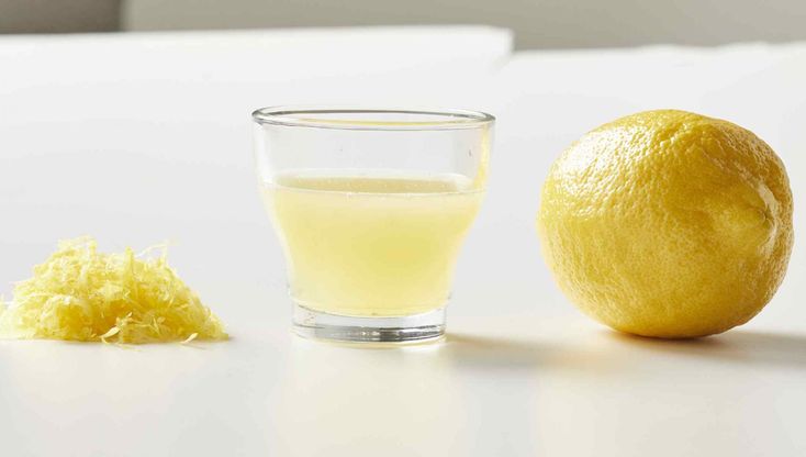 a glass of lemon juice next to an orange on a white counter top with the peel peeled off