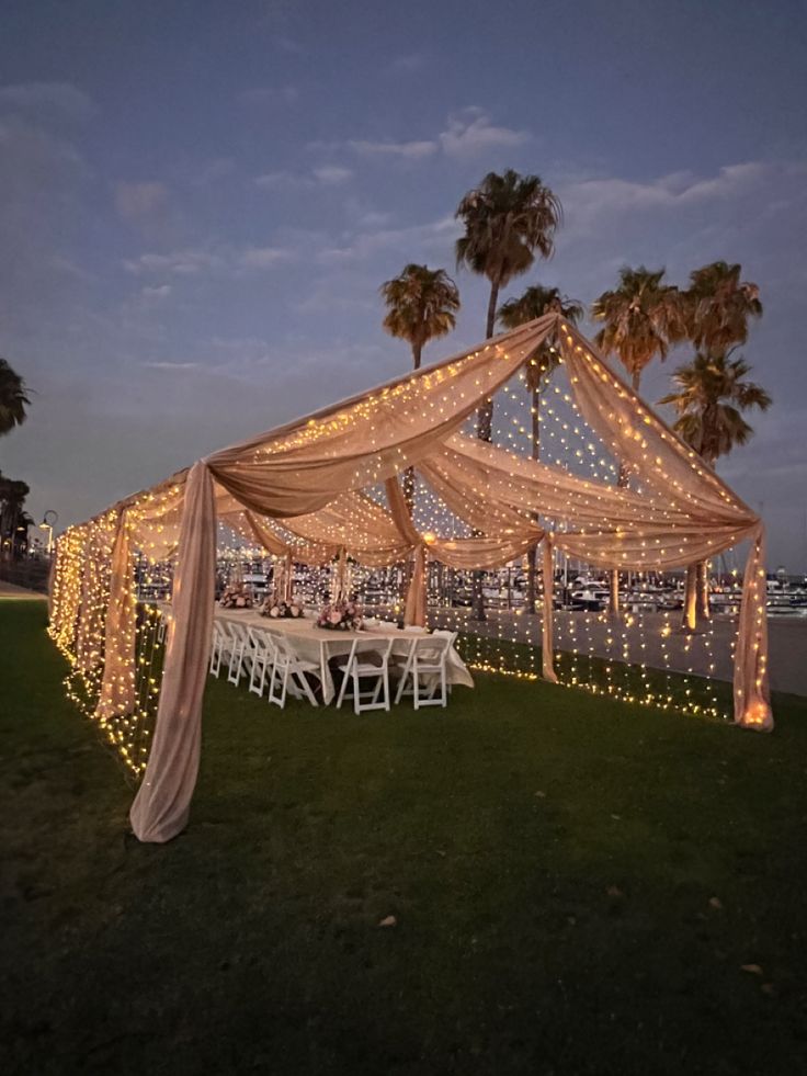 an outdoor wedding setup with lights and drapes on the grass by the water at night