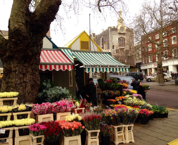 an outdoor market with flowers and plants on the sidewalk in front of some buildings near a tree