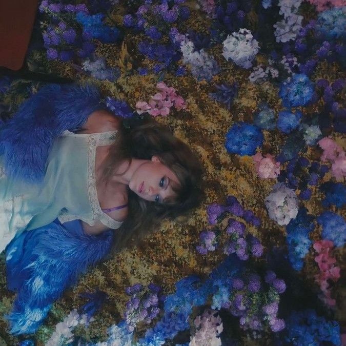 a woman laying on the ground surrounded by blue and purple flowers, with her hair blowing in the wind