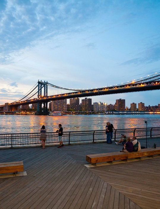 people are sitting on benches near the water with a bridge in the background at dusk