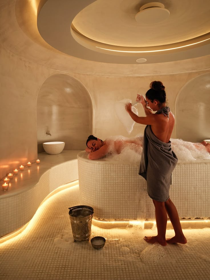 a man and woman in a spa room with candles on the walls, one person taking a bubble bath