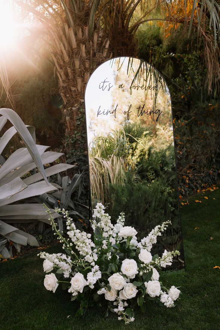 white flowers are sitting in front of a large mirror on the grass near some palm trees