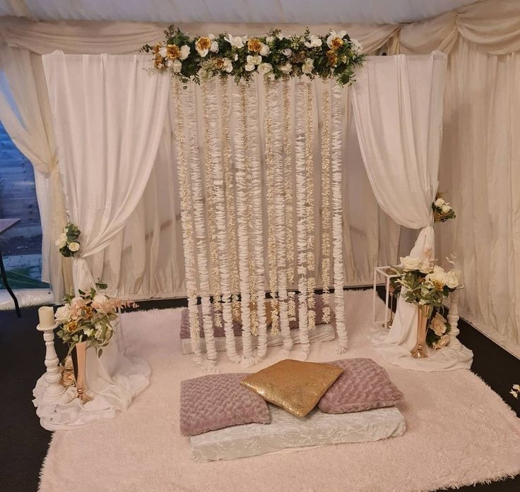 a wedding ceremony setup with white drapes and flowers on the wall, along with gold pillows