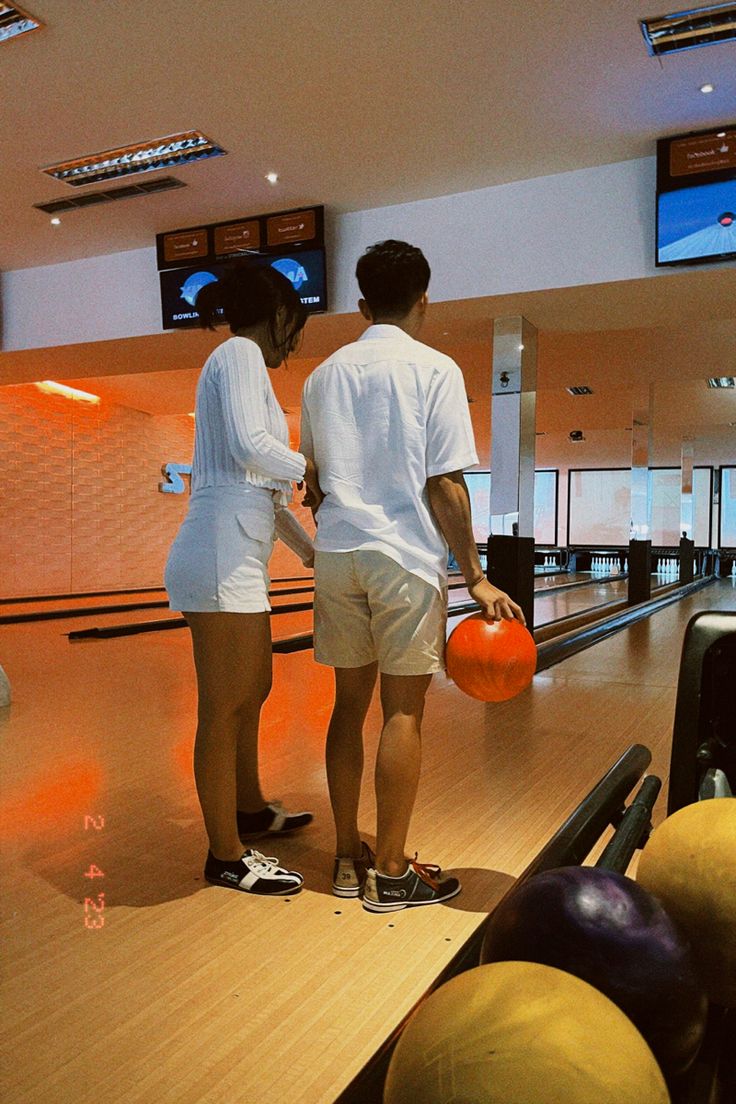 two people standing on a bowling alley holding orange balls