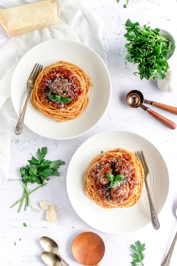 two white bowls filled with spaghetti and parsley on top of a table next to utensils