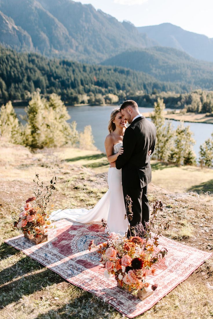 a bride and groom standing on a blanket in front of a lake with mountains behind them