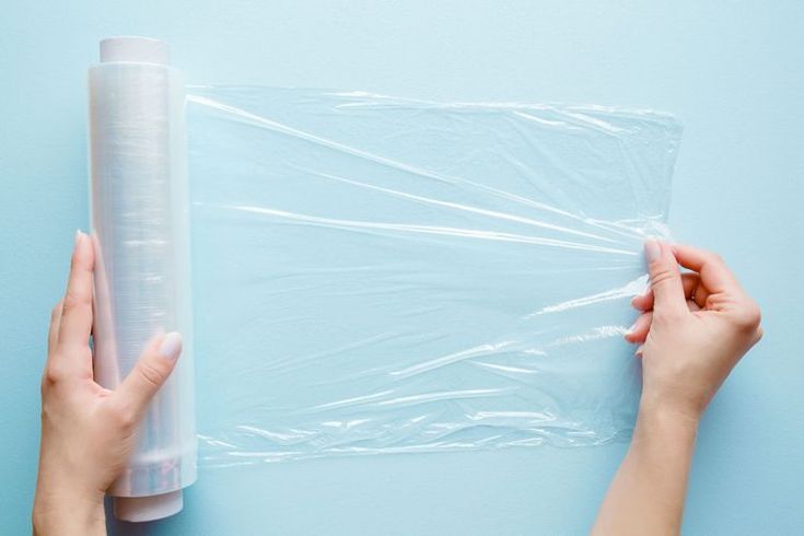 two hands holding a clear plastic bag on top of a blue surface next to a roll of toilet paper